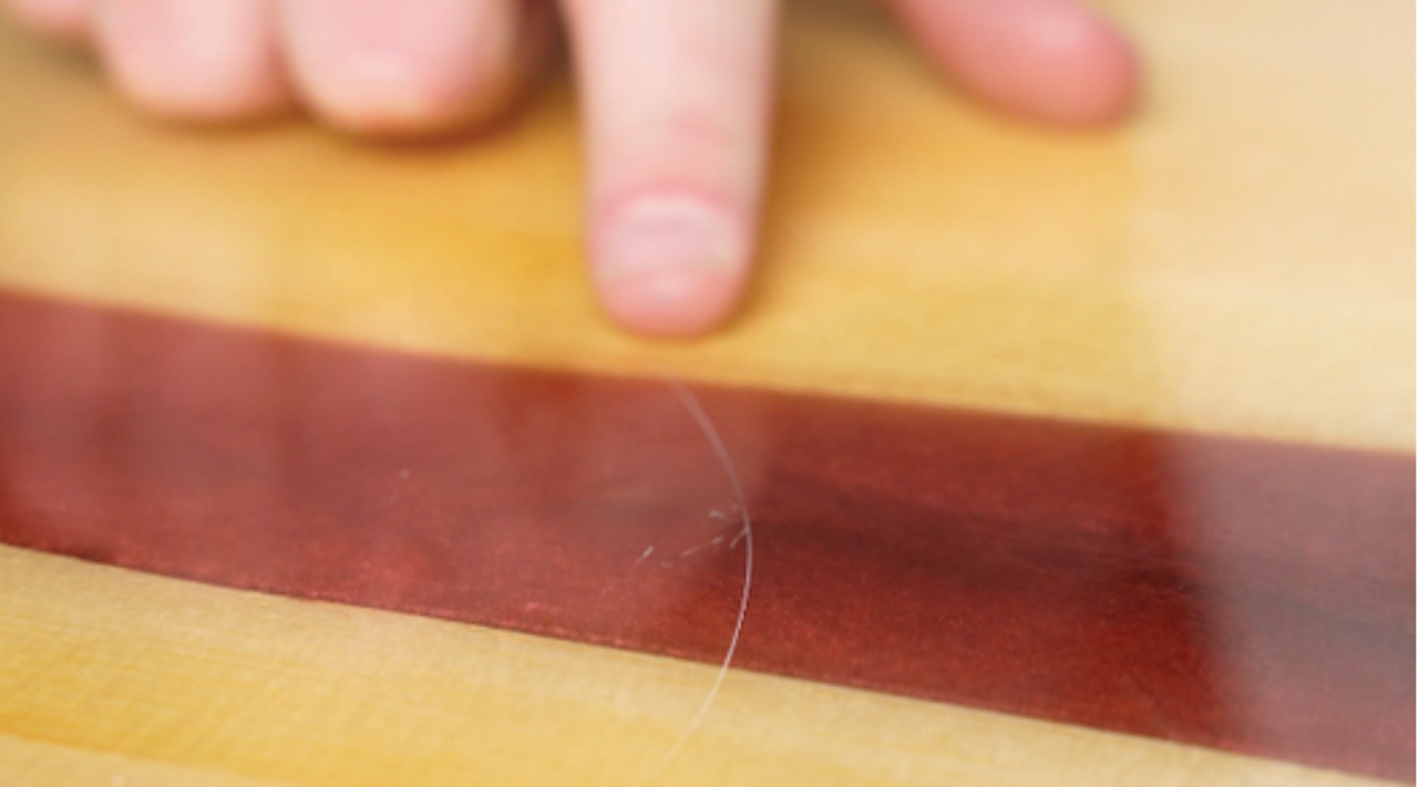 white hand pointing at a scratch in a wooden resin board with a red stripe down the middle to show how to sand epoxy resin