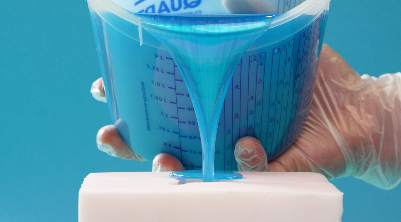 A person wearing gloves pours vibrant blue epoxy resin from a measuring cup onto a white surface, demonstrating the precise application and thickness that differentiates epoxy resin vs. other adhesives.