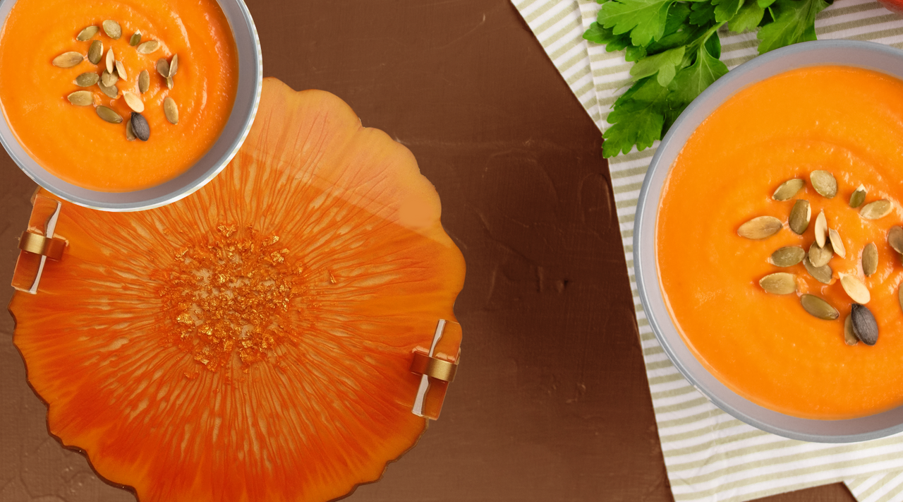 A vibrant orange resin tray with thin epoxy next to two bowls of creamy pumpkin soup garnished with pumpkin seeds, evocative of thin epoxy applications in its smooth texture and consistent color.