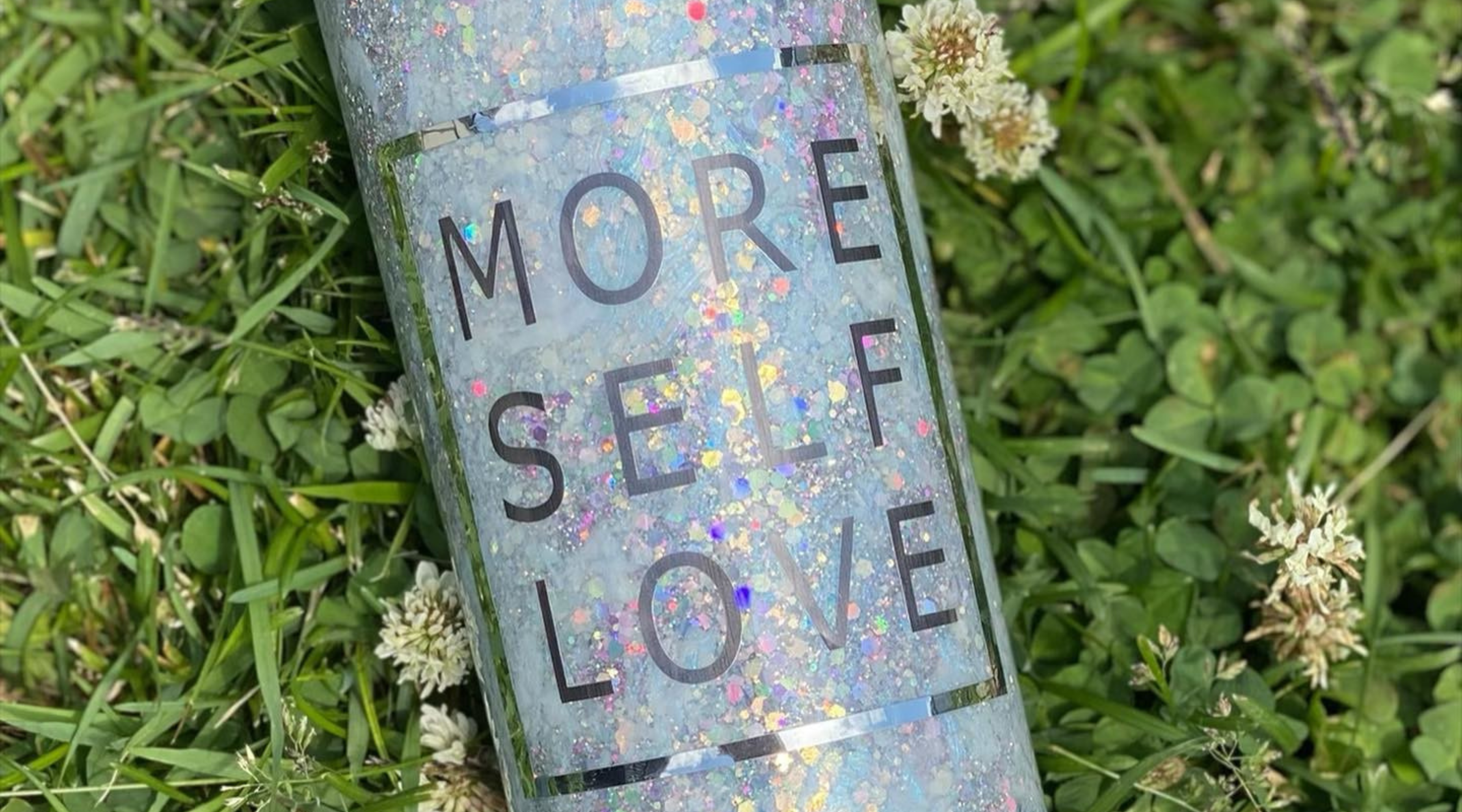 silver glitter tumbler with a "more self love" decal sitting in the grass with dandelions showing how to make tumblers